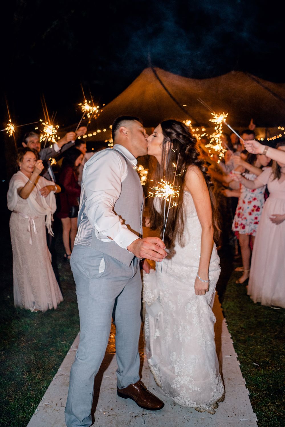 Couple shares romantic kiss under sparklers during outdoor tent wedding reception.