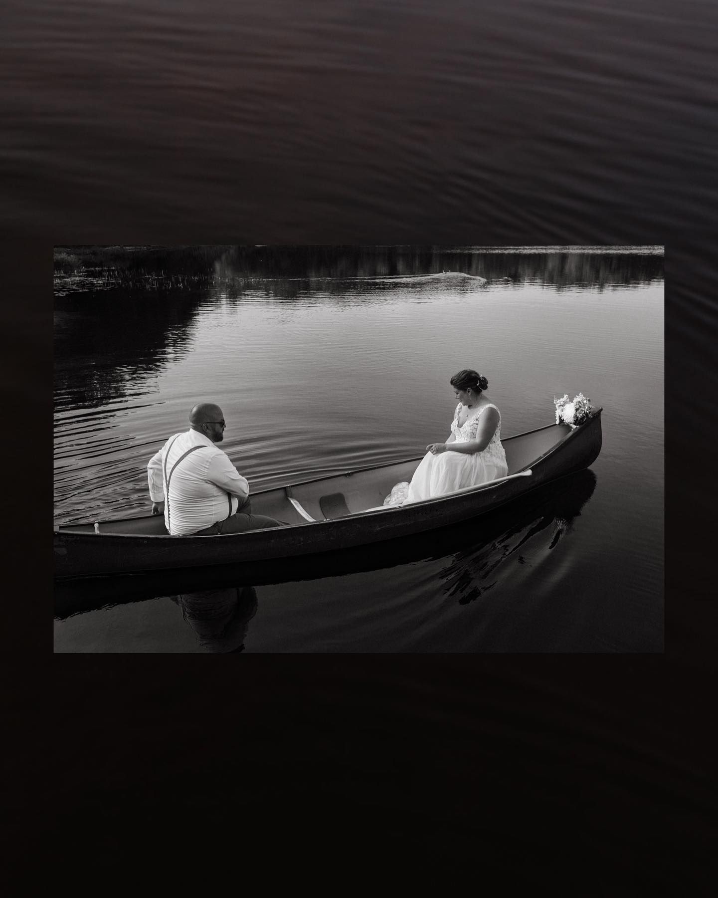 Black and white image of a couple in a rowboat on a serene lake during their wedding.