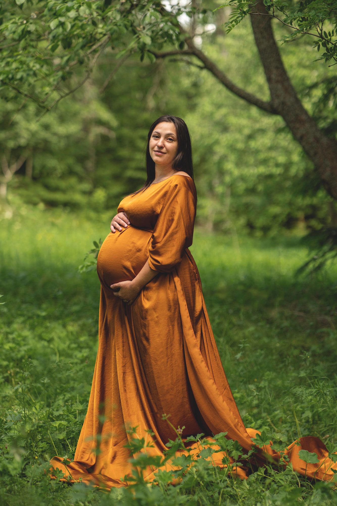 En gravid kvinna i rostorange boho klänning håller om sin gravidmage. Hon står i grönskan under ett träd.