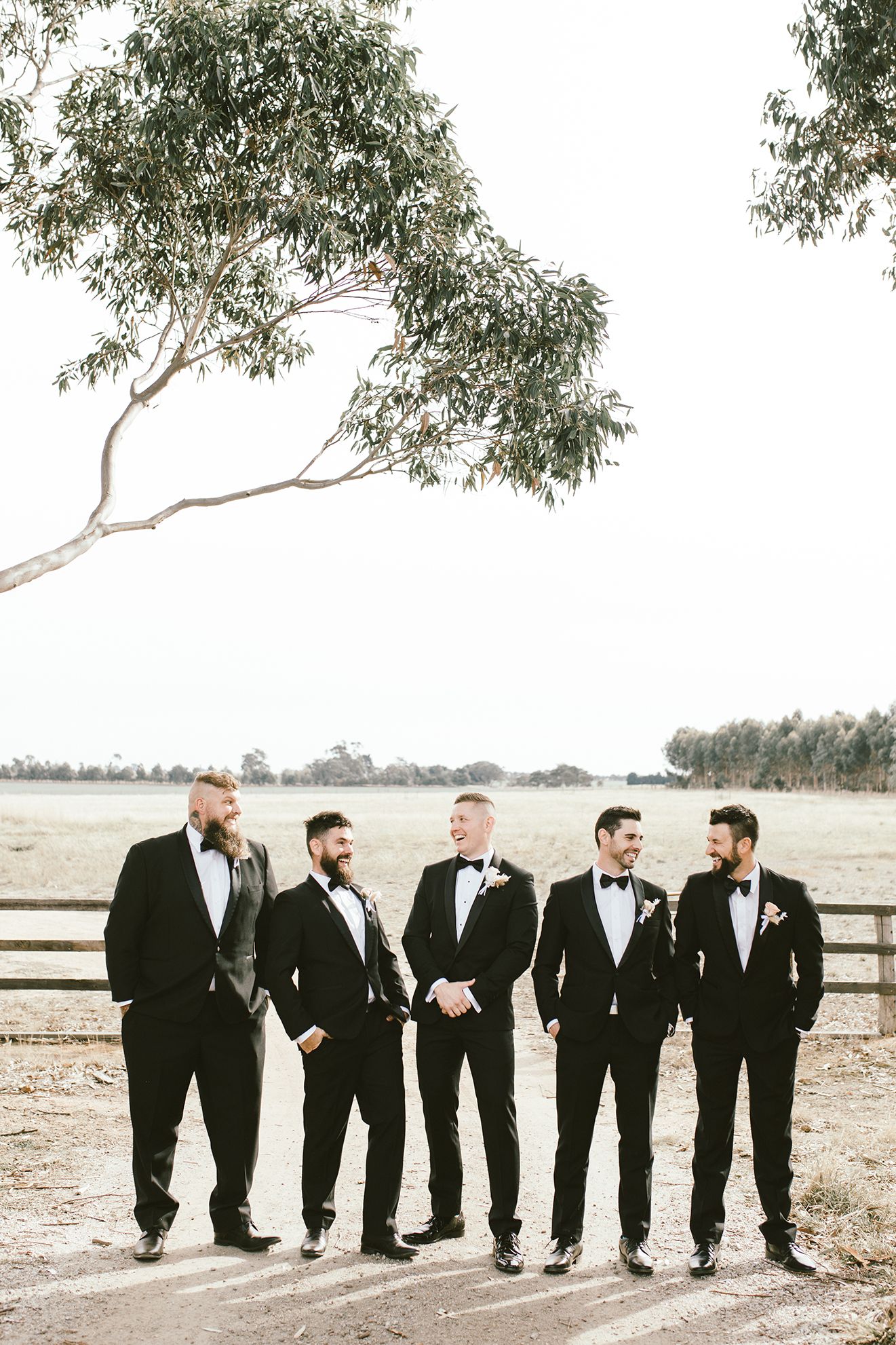 Groomsmen in classic black tuxedos stand together in rustic outdoor setting.