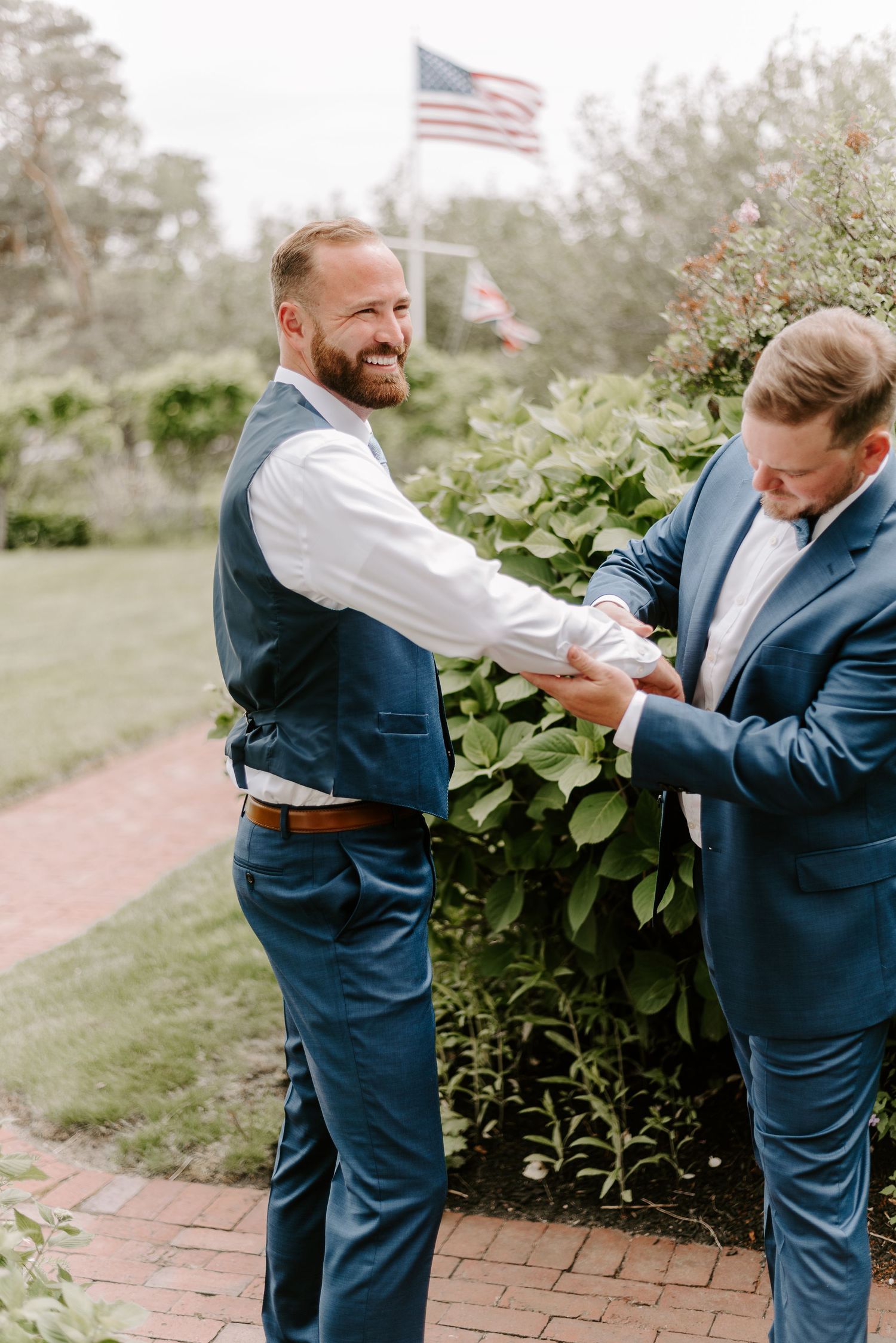 Two men in navy suits playfully adjusting each other's attire outdoors near an American flag.