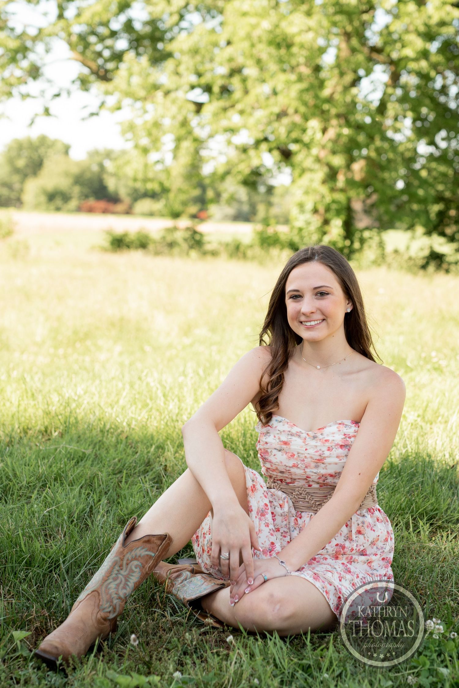 Summer portrait in floral dress and cowboy boots sitting in grassy field with barn background.
