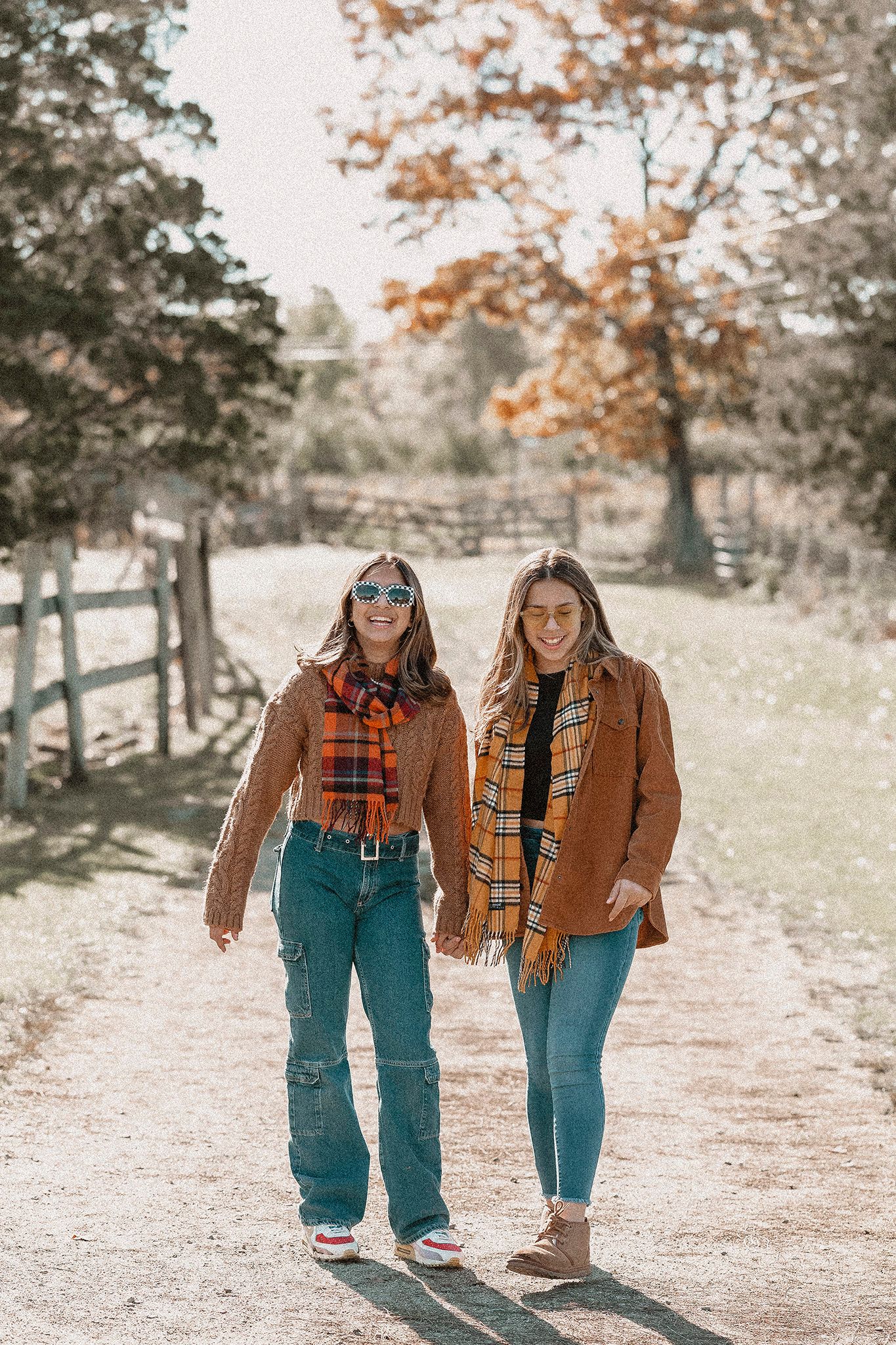 Two friends in matching brown jackets and plaid scarves walking along a country path in autumn.
