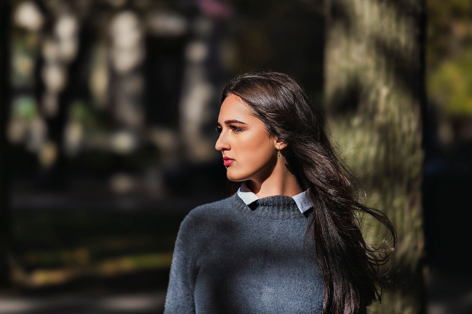 Side profile portrait outdoors wearing grey sweater with sunlit background.