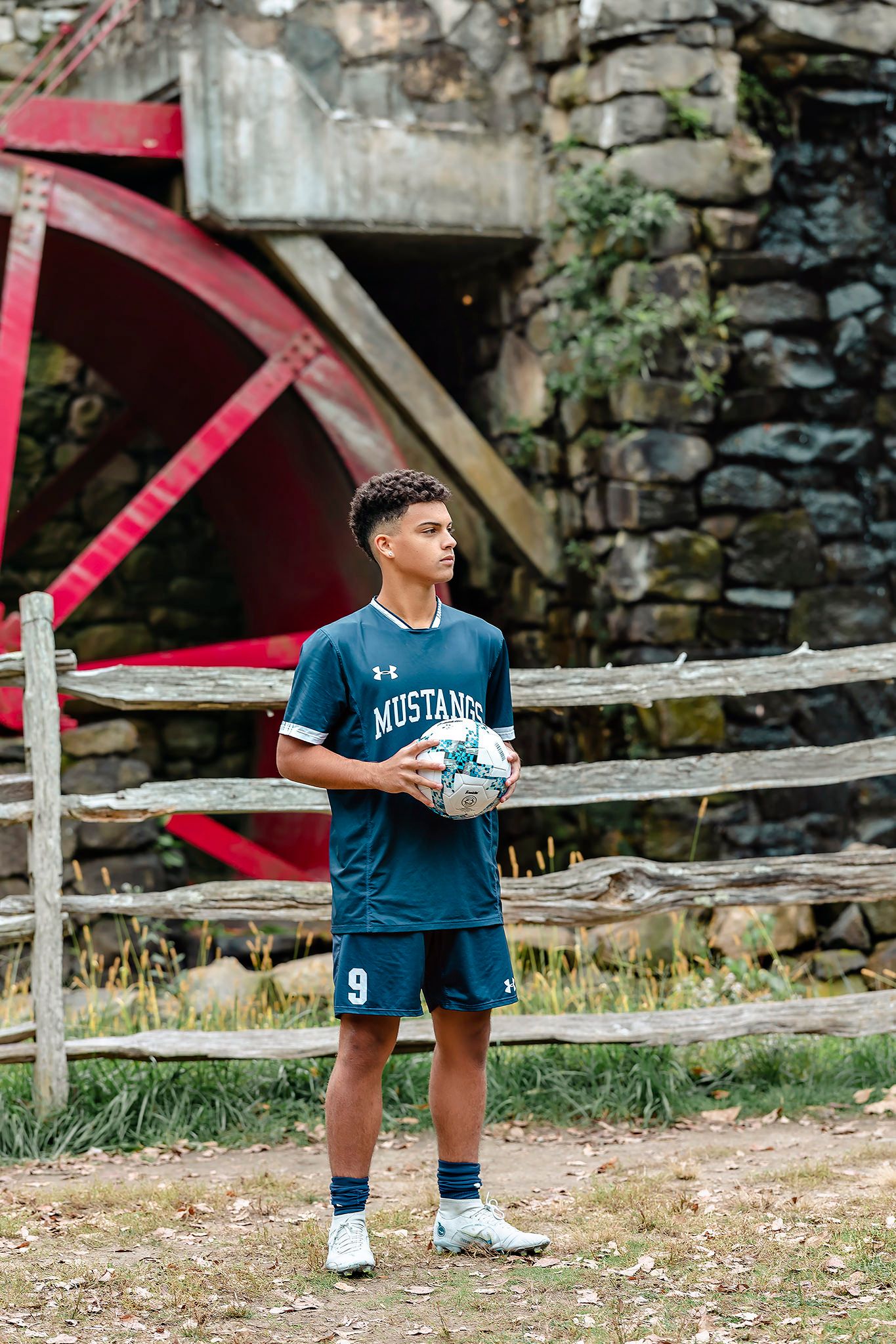 Young athlete in blue Mustangs uniform holding soccer ball at rustic outdoor location.