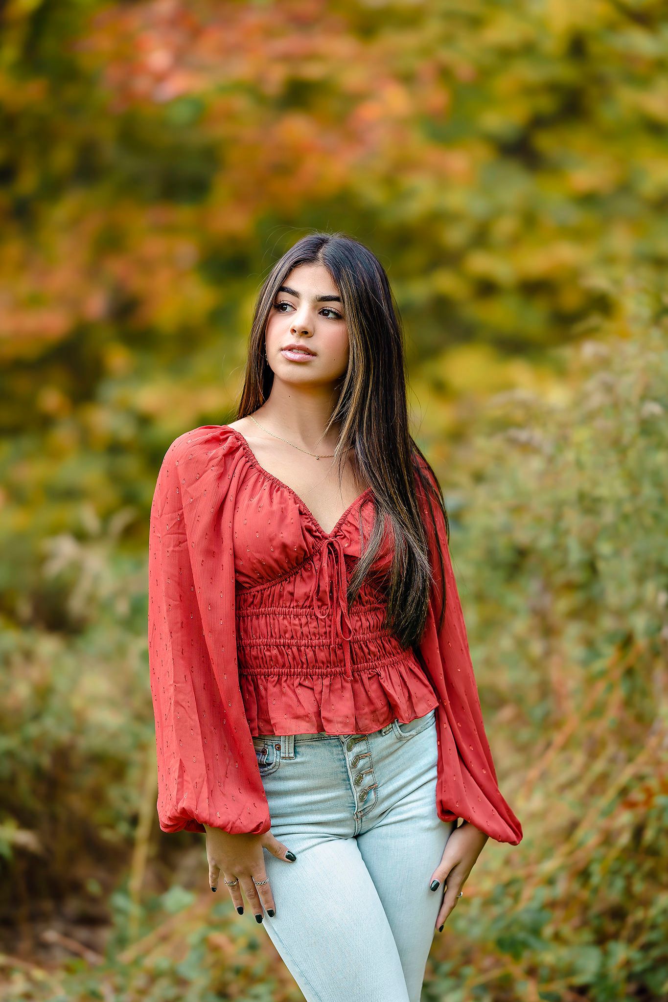 Young person in a red peasant blouse and light jeans poses outdoors with autumn foliage in the background.