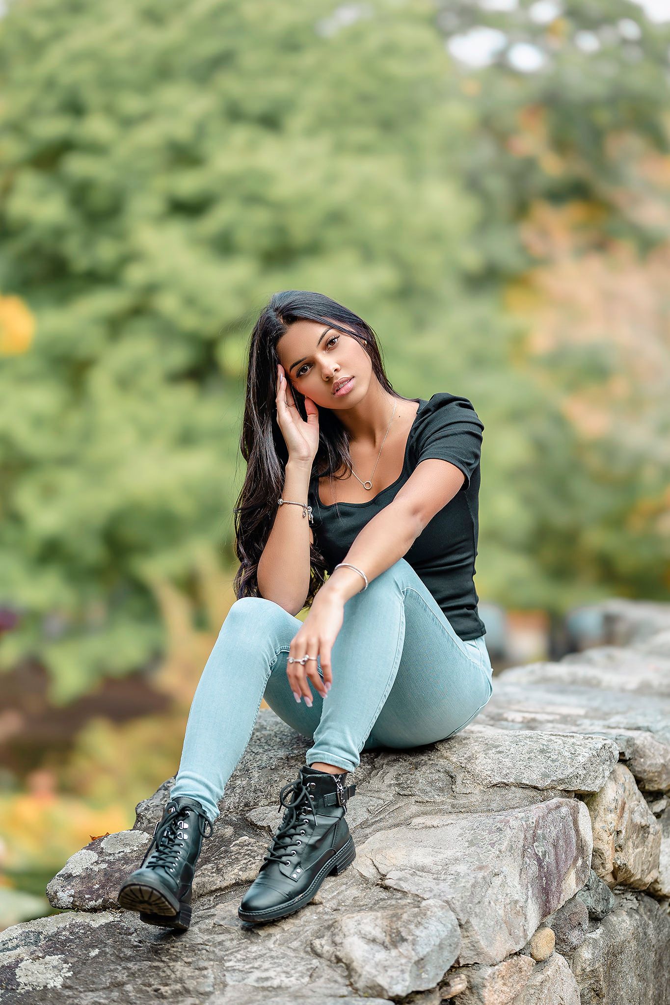 A young woman in casual attire sits relaxed on a rocky outcrop in a natural outdoor setting with green foliage behind her.