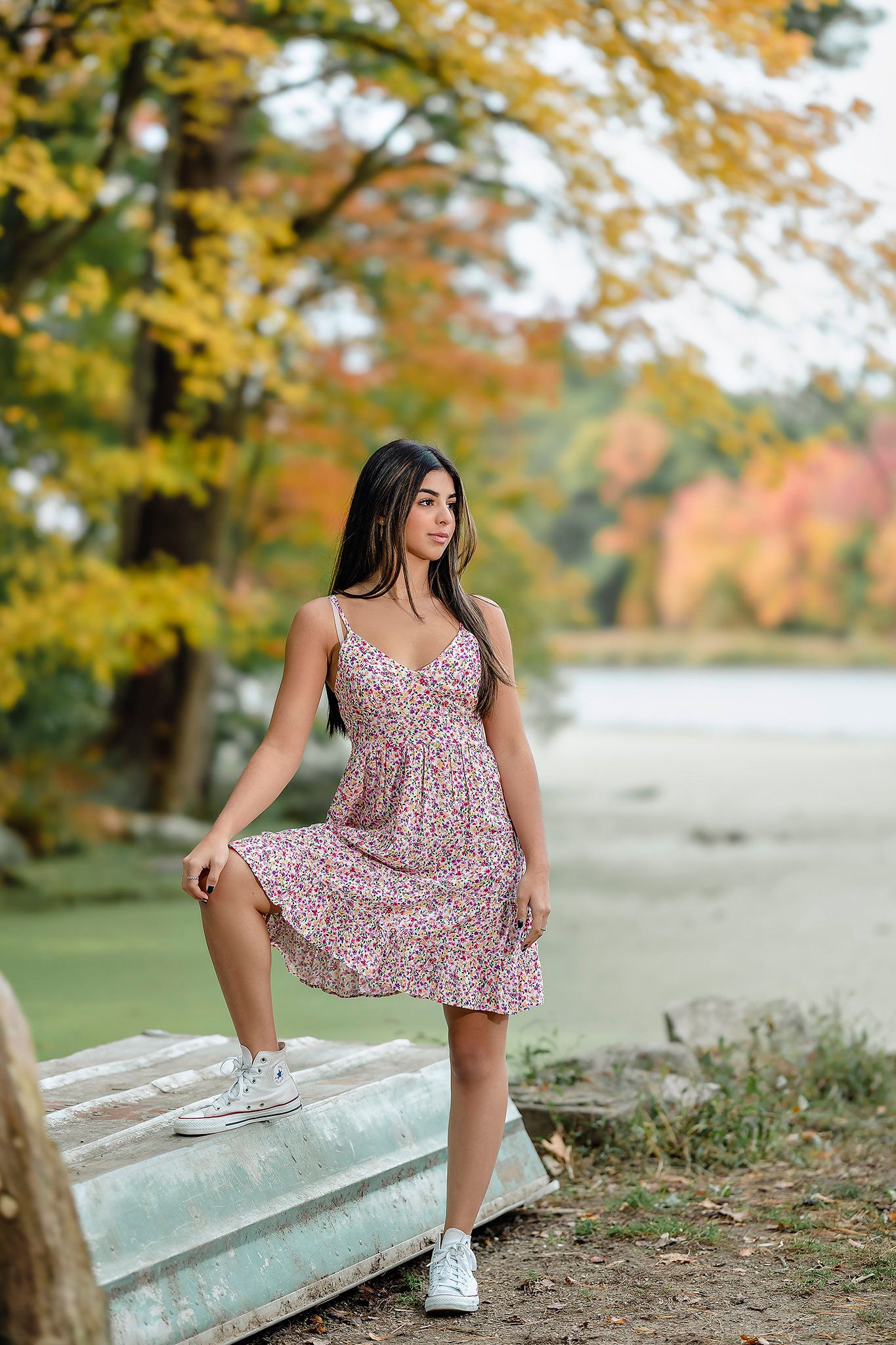 Person in a floral dress poses in front of autumn trees and a lake during an outdoor portrait session.