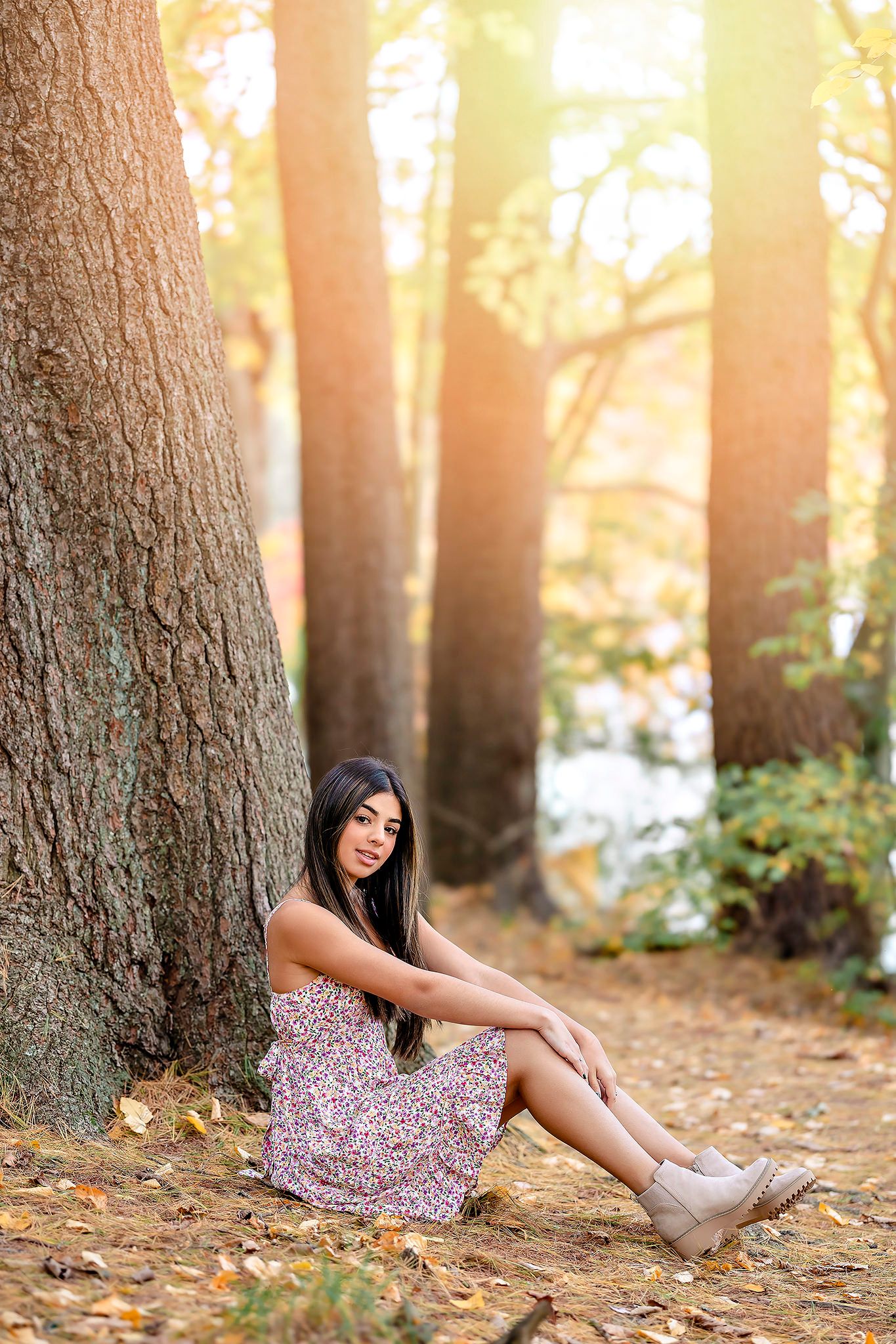 Person in a floral dress sits among fallen autumn leaves beneath tall trees at sunset with warm golden light filtering through.