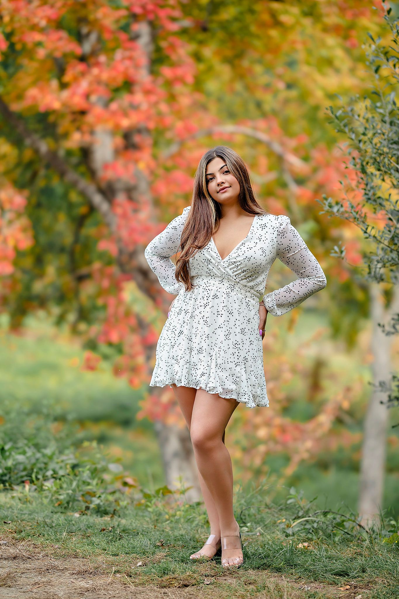 A woman in a white floral dress poses gracefully in front of autumn foliage with vibrant red and orange leaves.
