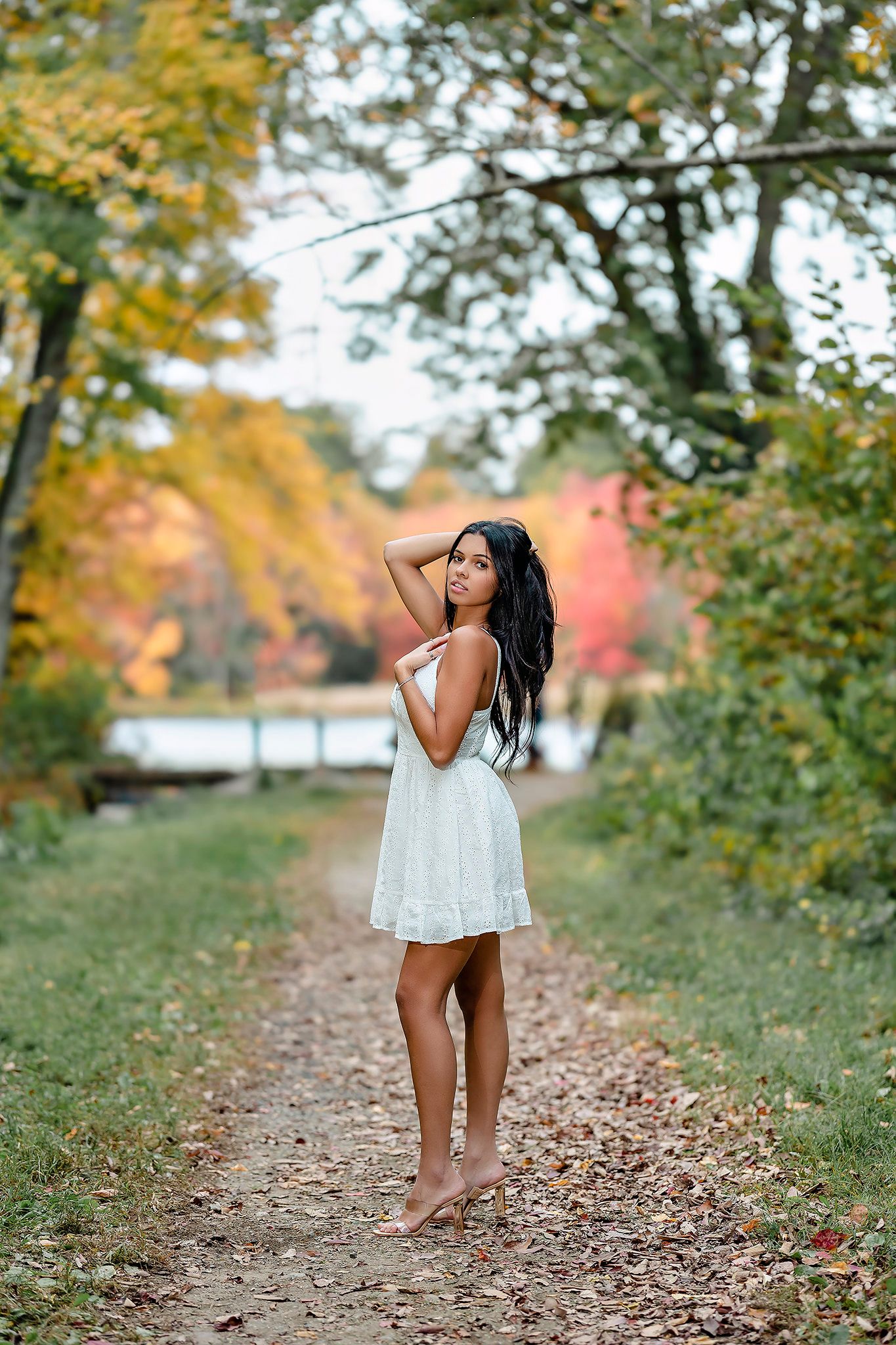 A barefoot woman in a white dress stands on a autumn leaf-covered path surrounded by colorful fall foliage.