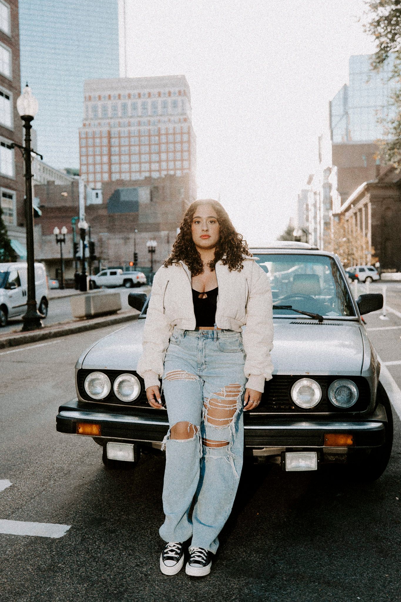 Person wearing white jacket and ripped jeans poses against vintage black car on urban street.