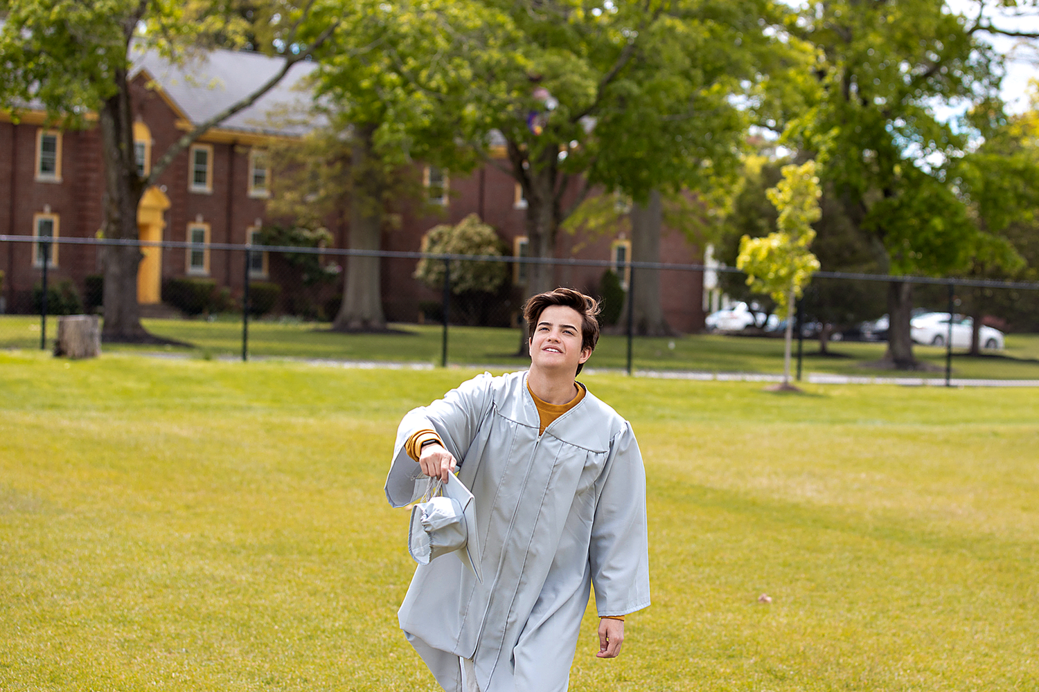 Graduate in light gray gown celebrates on sunny campus lawn.