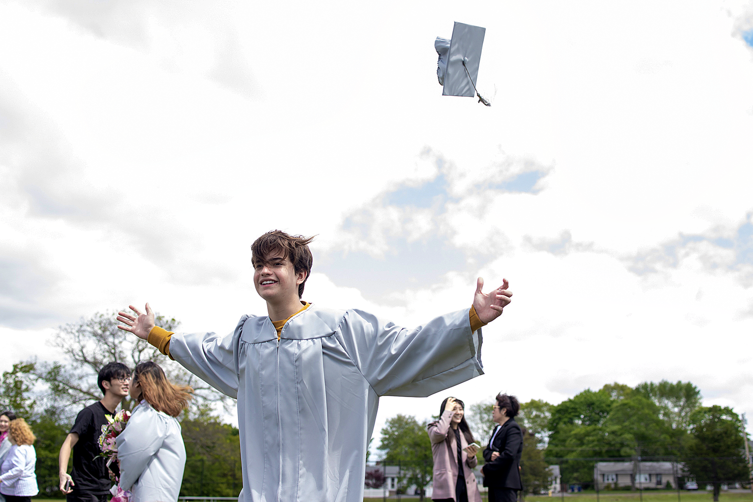 Graduate in light blue gown tossing cap in air during outdoor celebration.