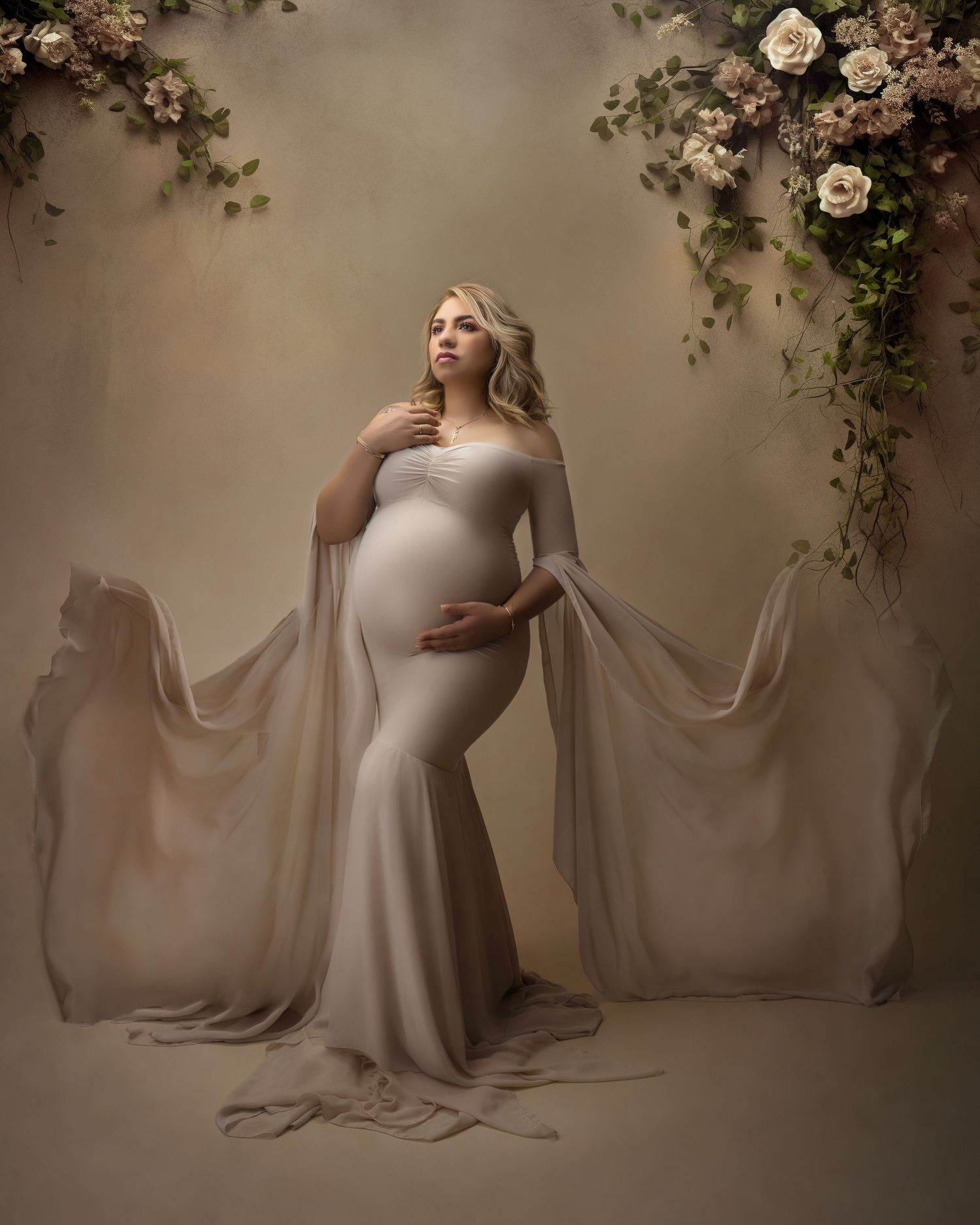 Elegant maternity portrait with flowing white dress and floral arch backdrop in dreamy lighting.