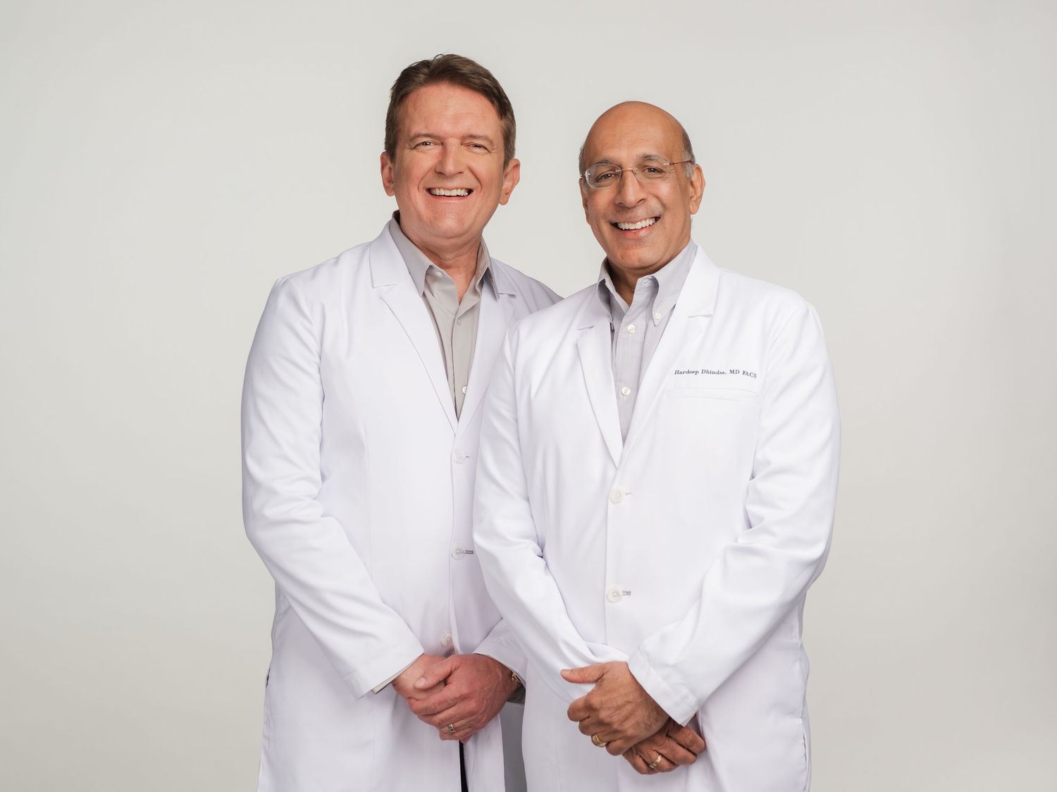 Two healthcare professionals in white medical coats stand together smiling against a light gray background.