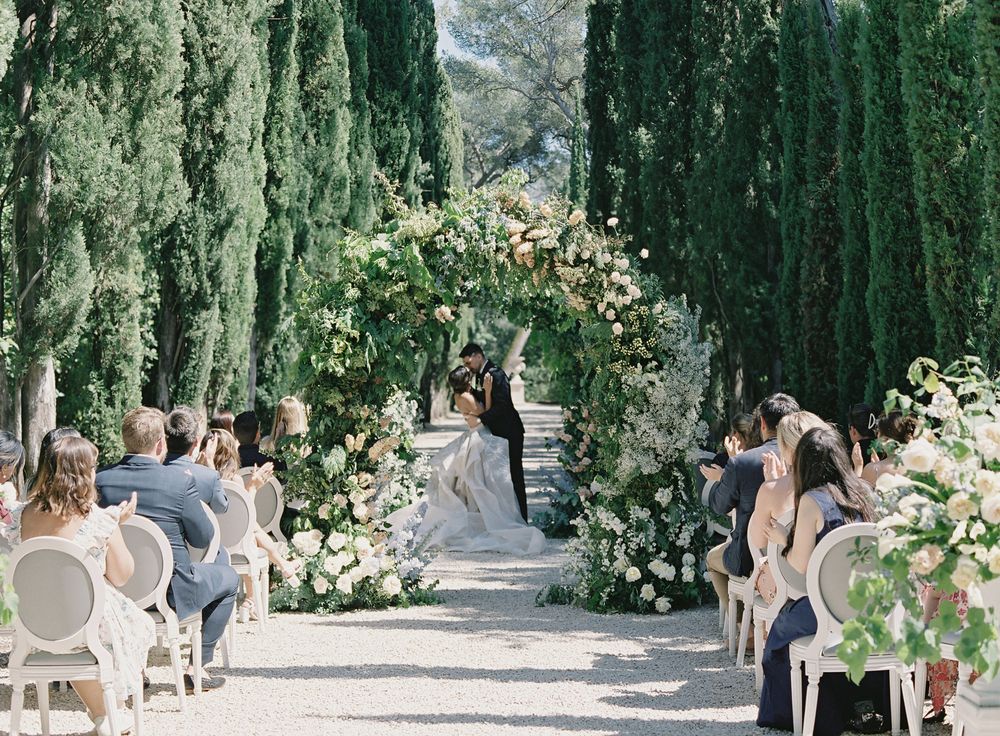 Romantic outdoor wedding ceremony with floral arch and white chairs set up between cypress trees.