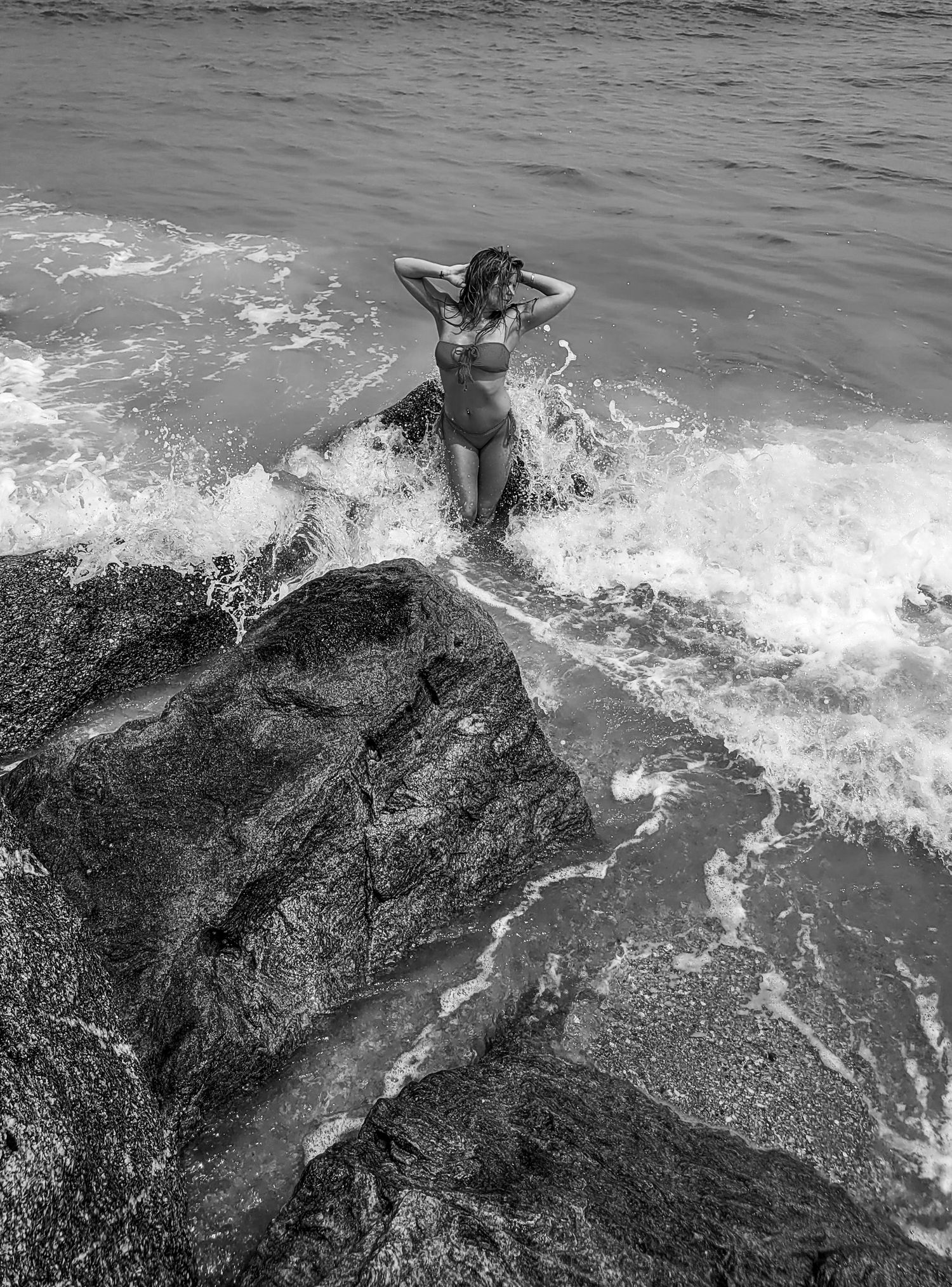 Black and white photo of person on rocky shore with crashing waves.