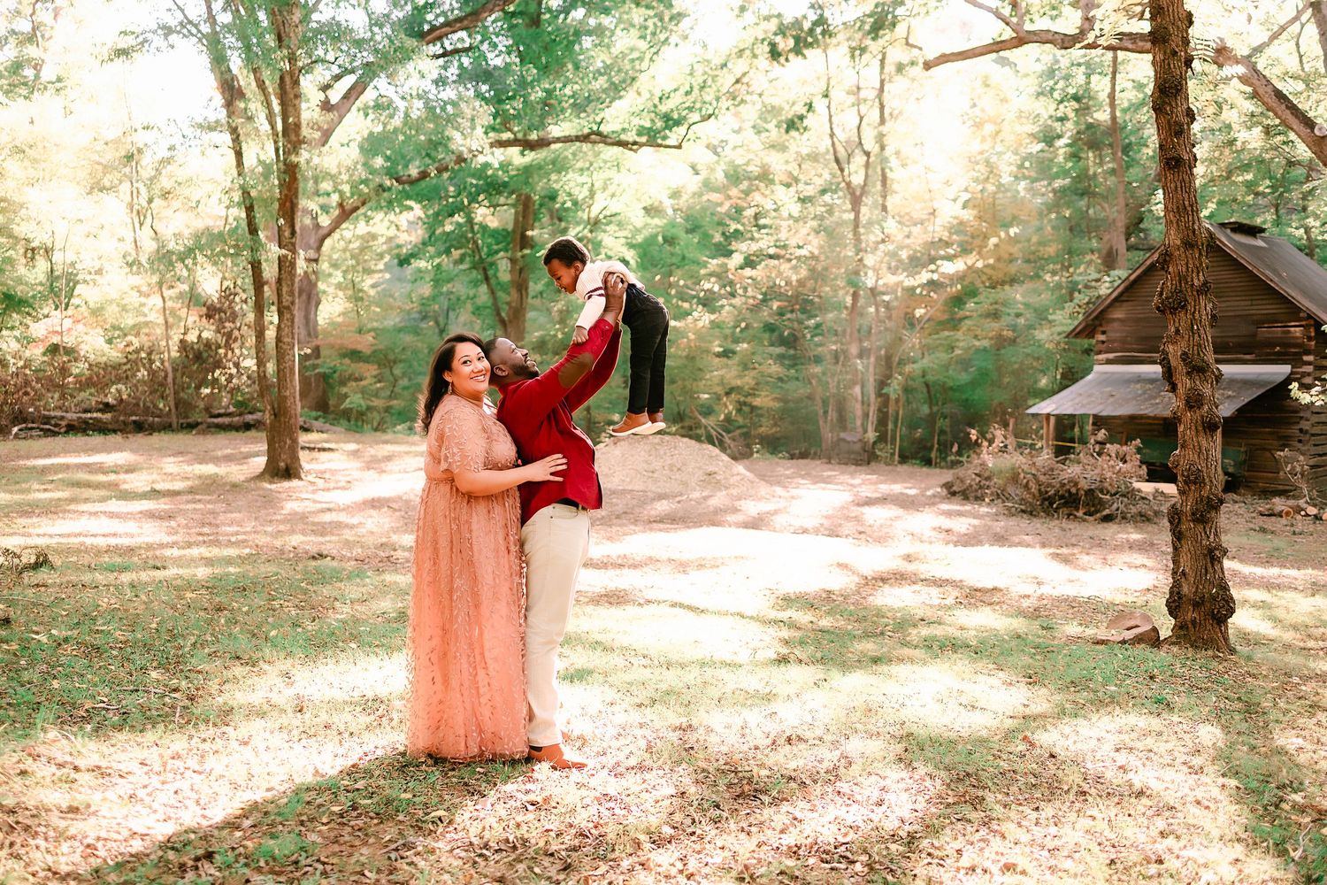 Couple posing playfully in a sunlit forest, woman in pink gown lifting man in burgundy suit near a rustic cabin.