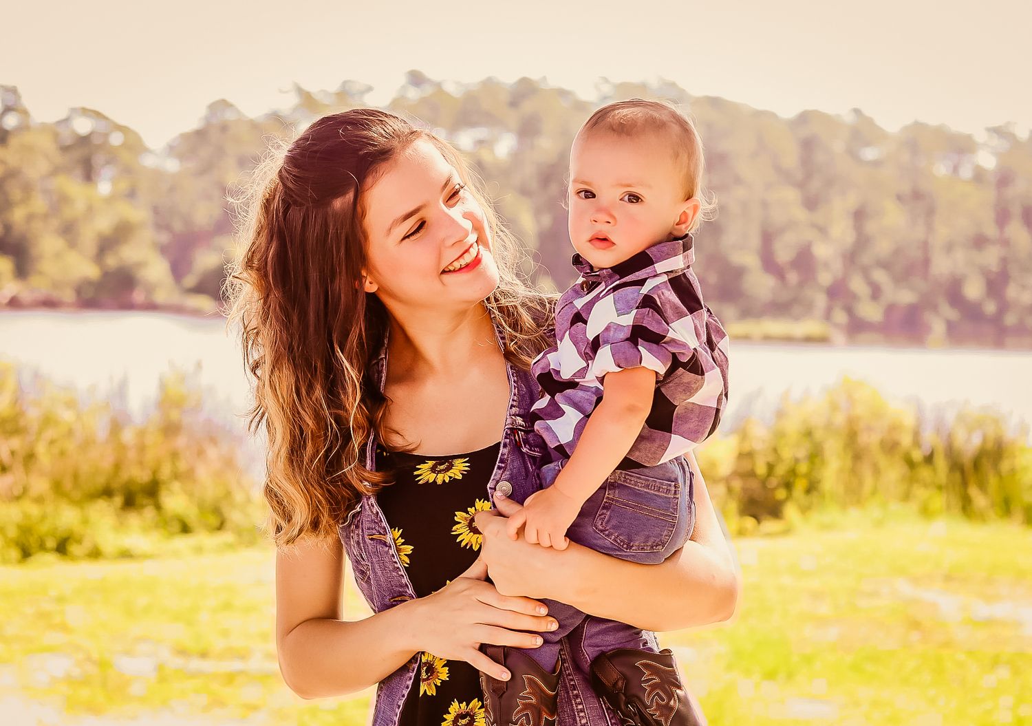 Young mother smiling and holding baby boy outdoors near a lake on a sunny day.