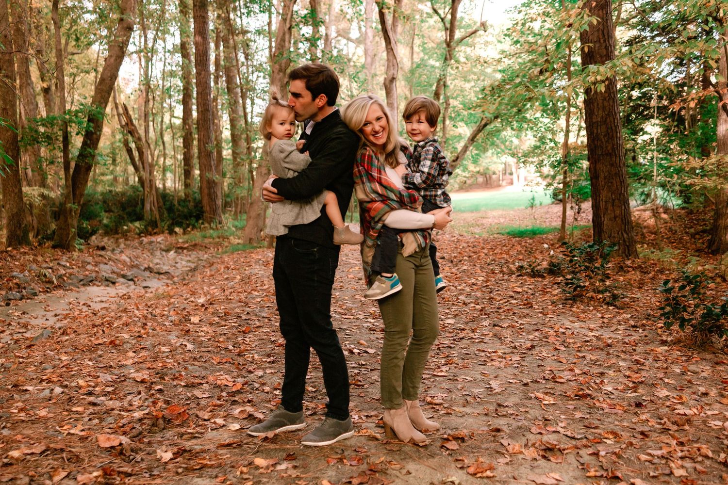 Family of four with two young children standing on a leaf-covered path in an autumn woodland setting.