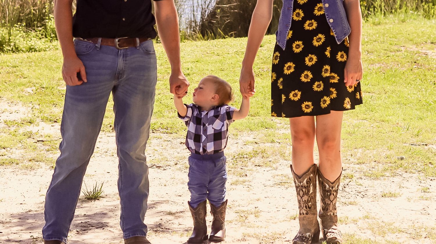 Family of three with parents holding toddler hands outdoors on sandy path.