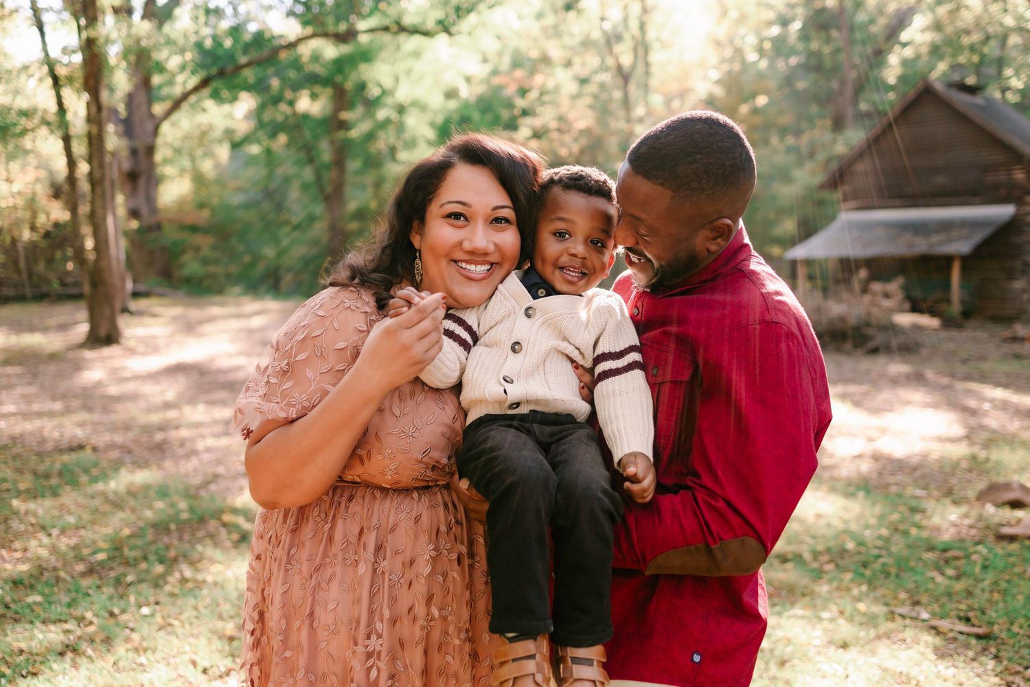 Happy interracial family smiling outdoors in autumn forest, parents holding young toddler son between them.