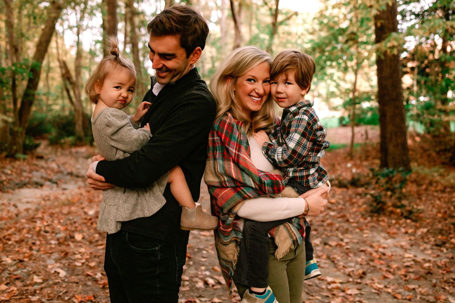 Smiling couple each holding a young child while posing back to back in a colorful autumn forest.