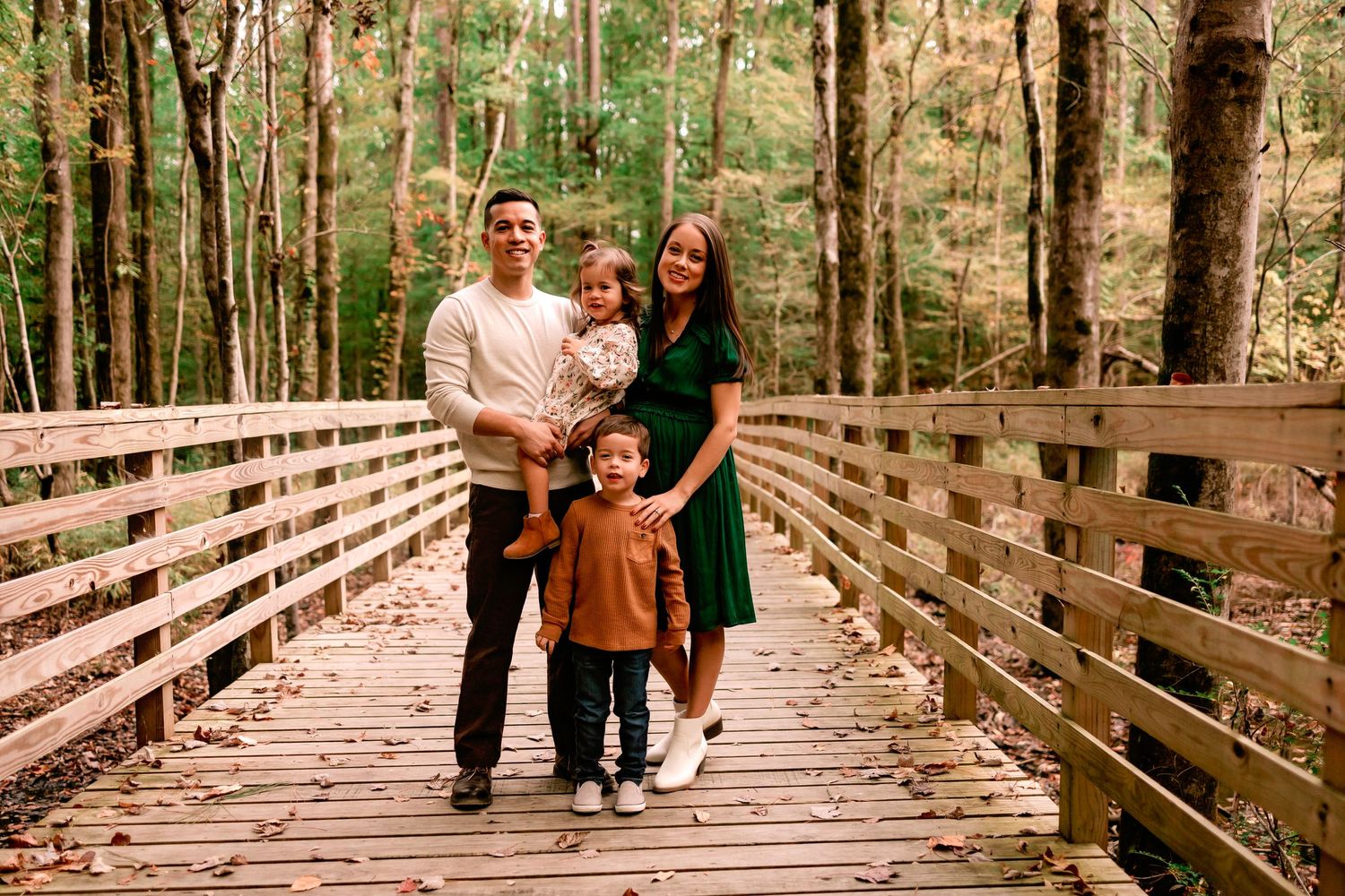 Family of four posing on a wooden bridge surrounded by colorful fall foliage in a forest setting.