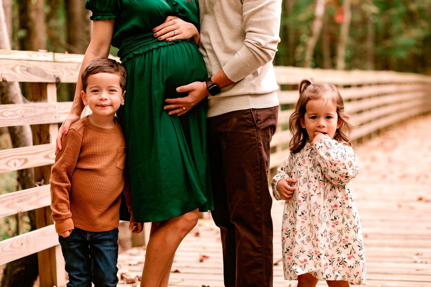 Pregnant mother in green dress with husband and two young children posing on a wooden bridge in fall.