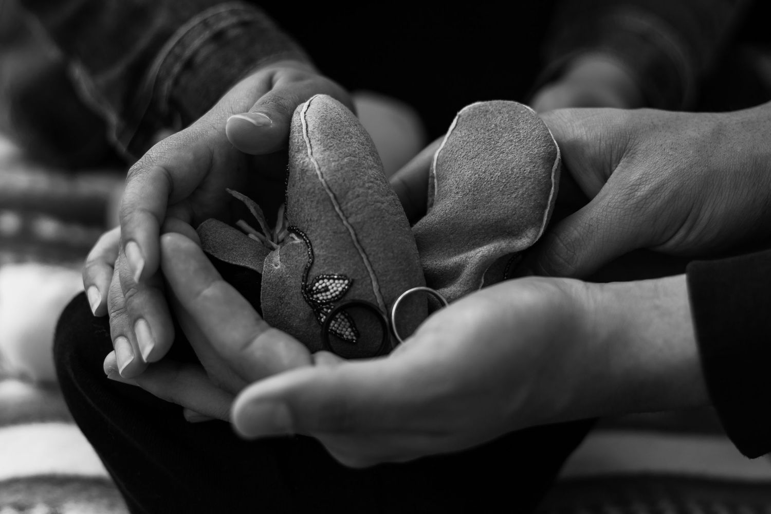 Black and white close-up of two pairs of hands holding tiny baby shoes with a ring.