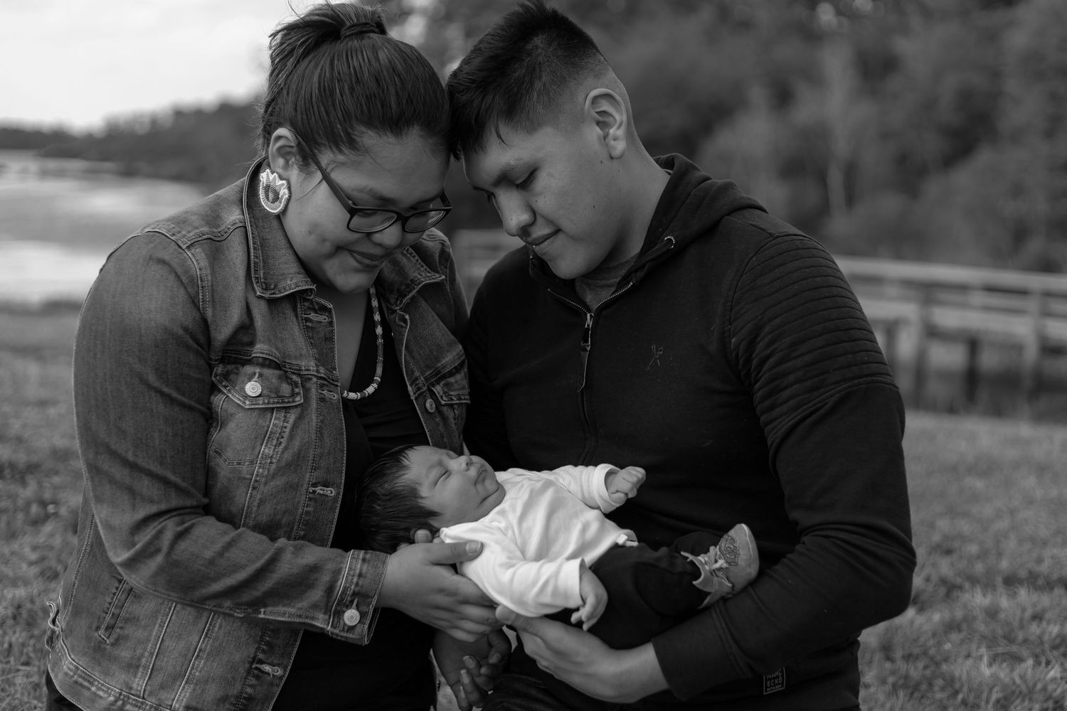 Black and white photo of a couple tenderly holding their newborn baby outdoors near a lake.