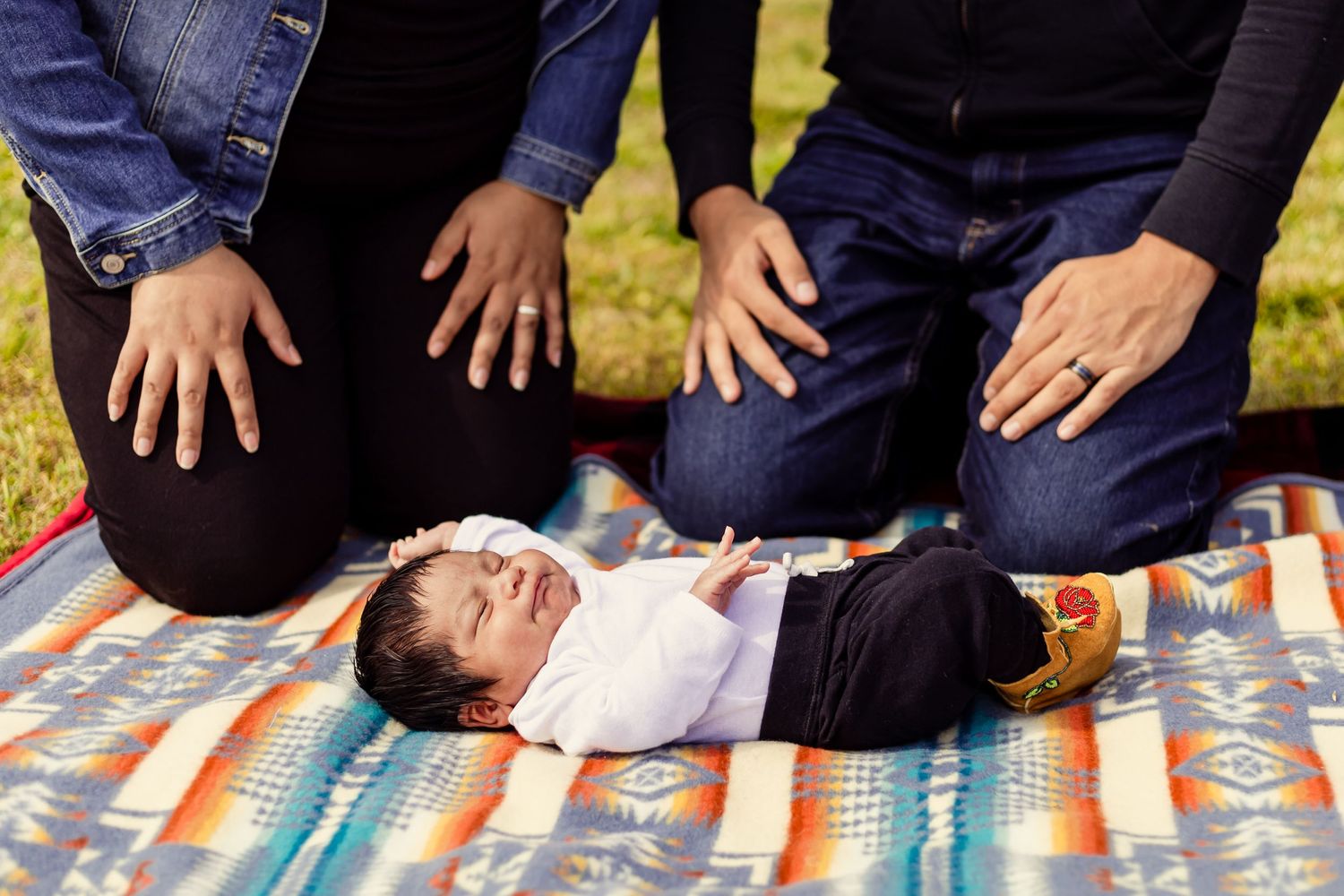 Newborn baby lying on colorful Pendleton blanket with parents kneeling beside, gently placing hands near infant outdoors.