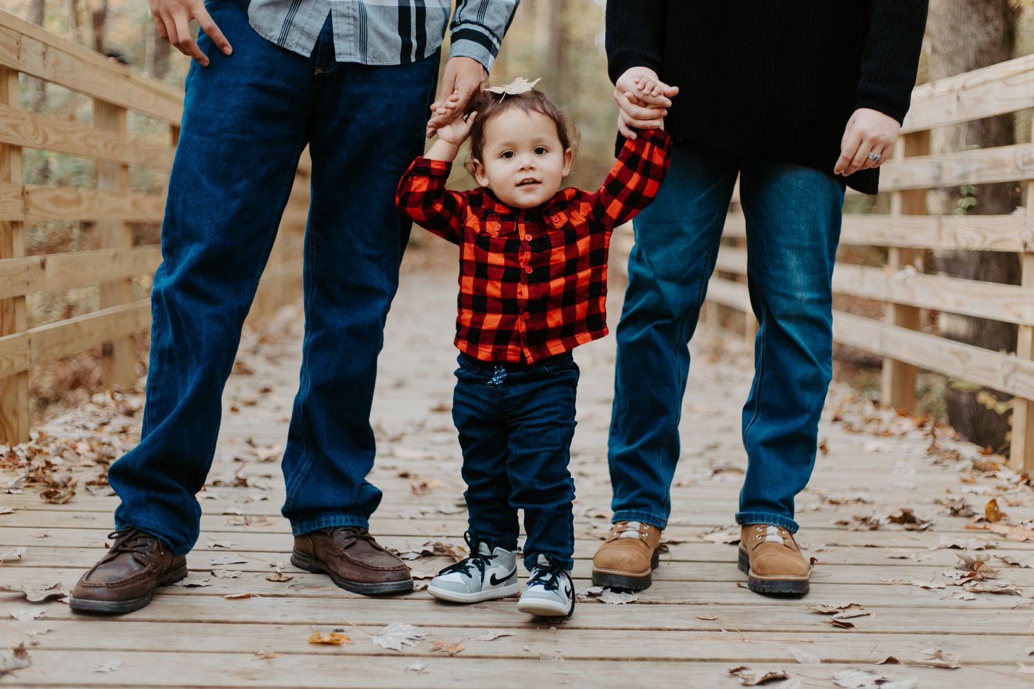 Toddler in red plaid shirt holding hands with two adults walking on a wooden bridge.