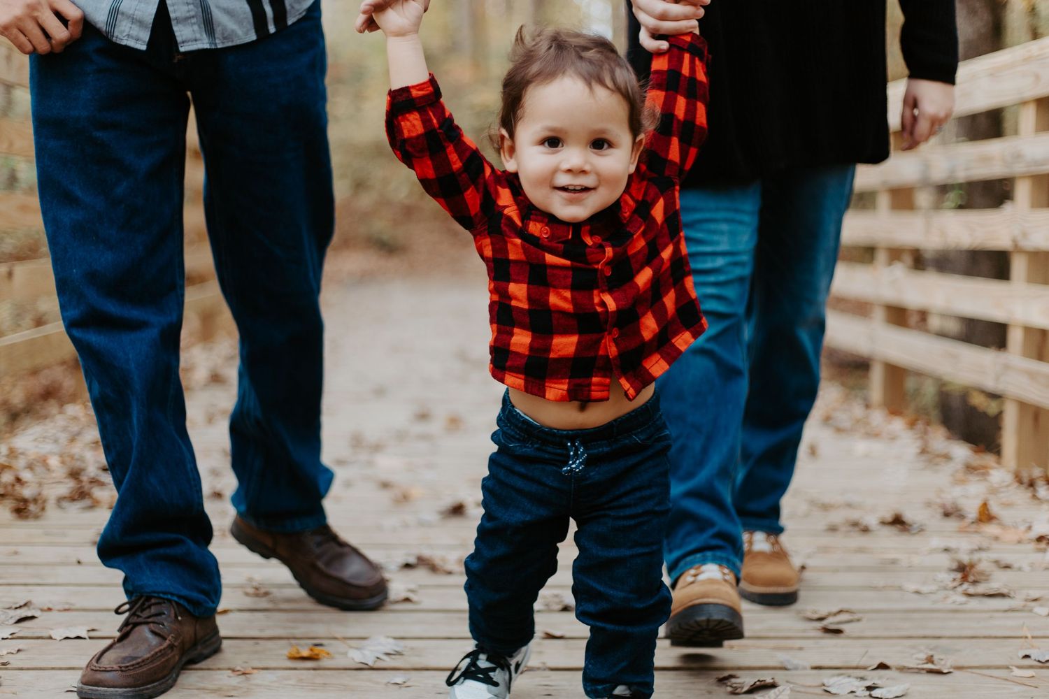 Smiling toddler in red plaid shirt walking on a wooden bridge with arms raised.