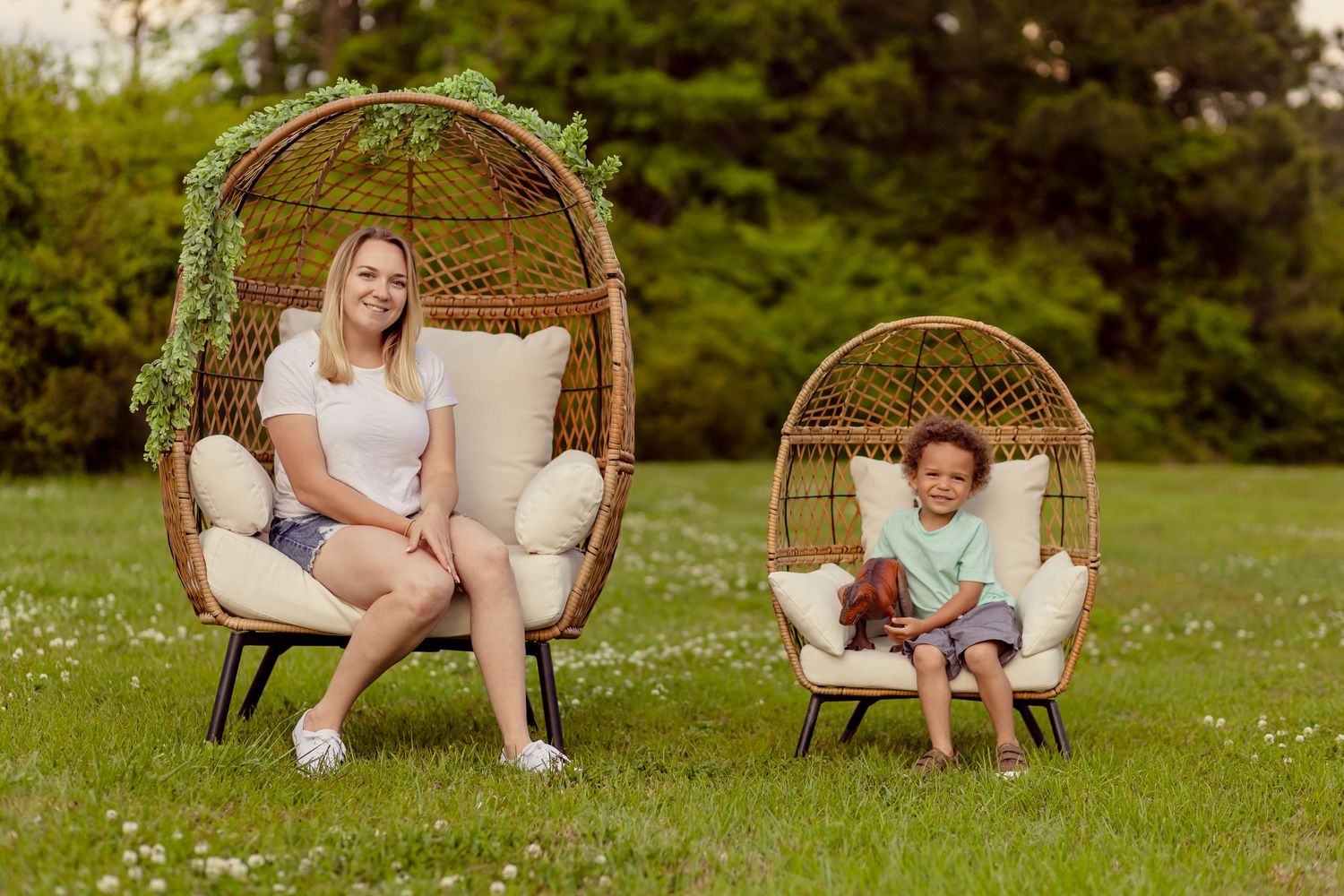 Blonde woman and young curly-haired child sitting in matching wicker egg chairs on green grass.