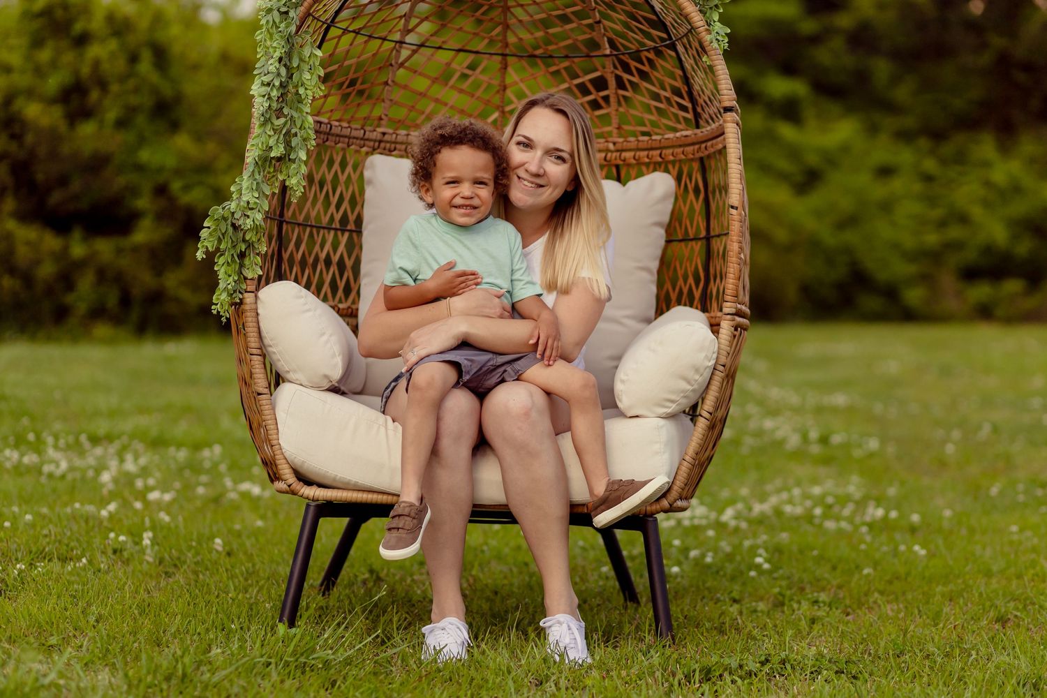 Smiling blonde woman hugging a young curly-haired child in a large wicker egg chair outdoors.