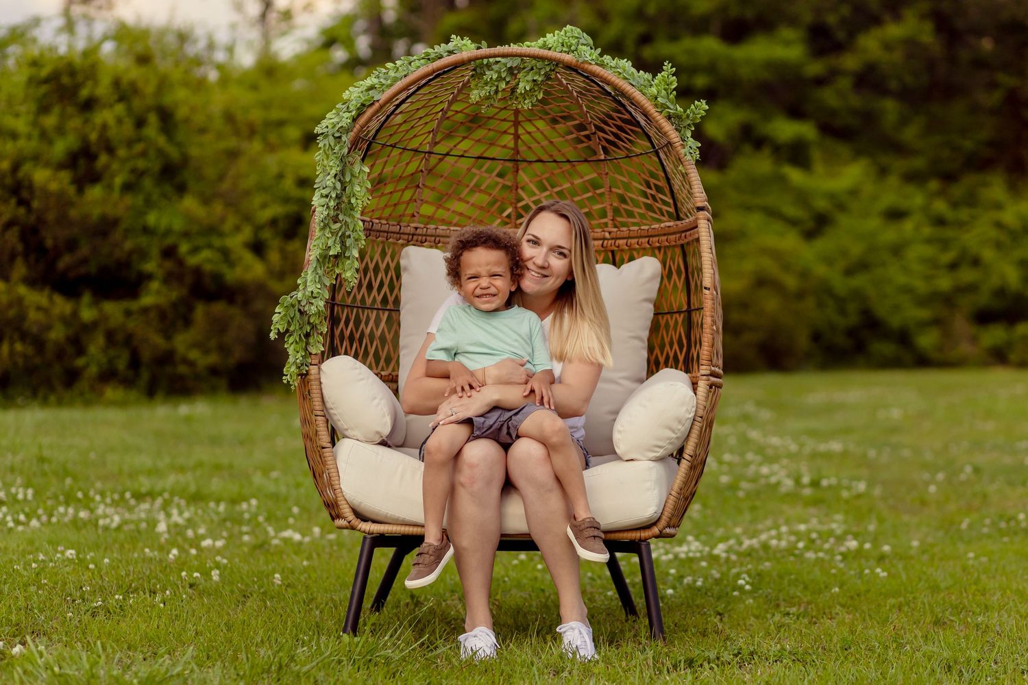 Happy blonde woman and young child posing together in a decorative wicker egg chair on grass.