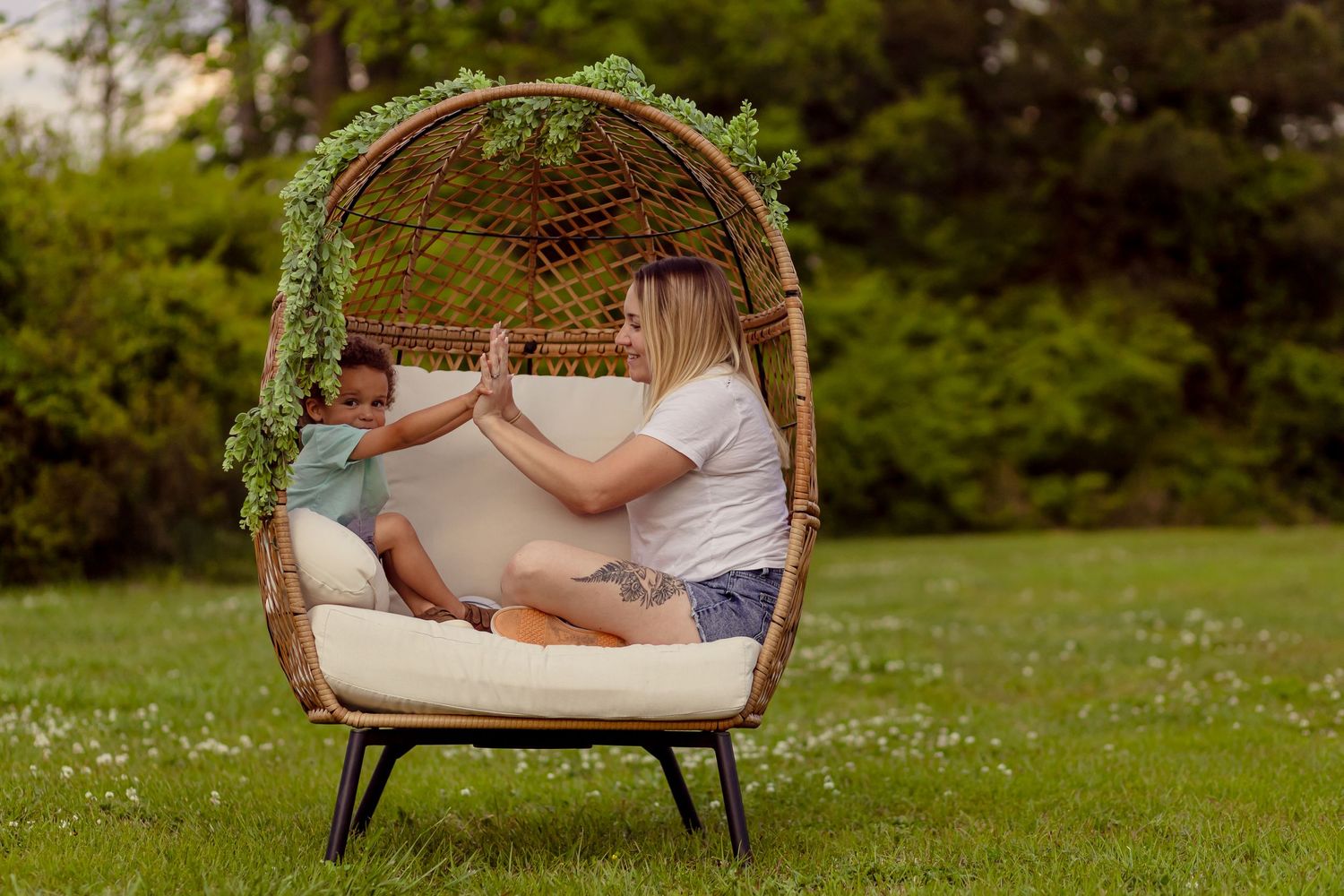 Blonde woman and young child playing patty-cake together inside a large wicker egg chair outdoors.