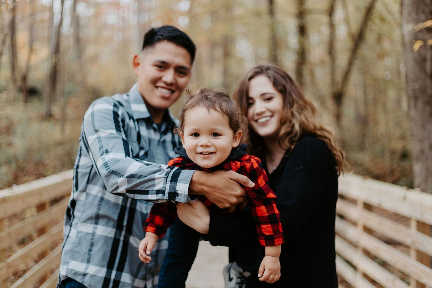 Smiling parents holding their toddler in a red plaid shirt on an autumn wooden bridge.