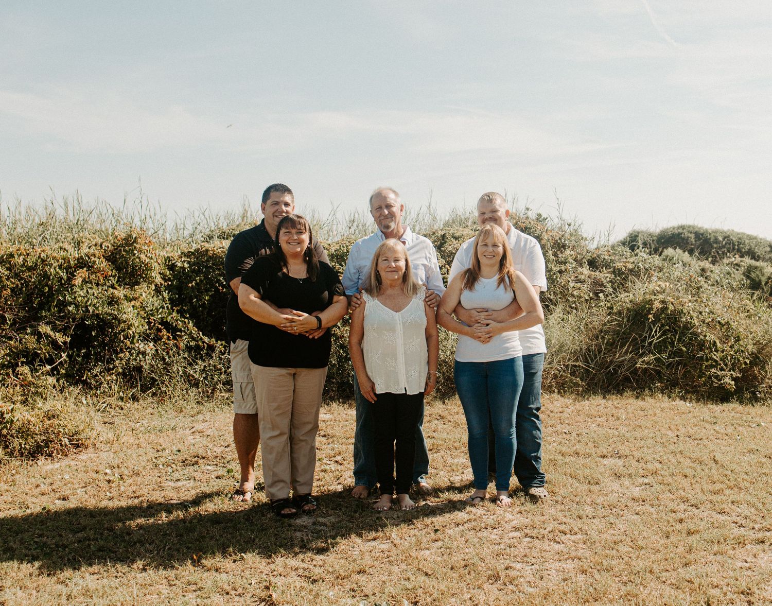 Group of six adults posing together outdoors in a grassy field with a bright sky behind them.
