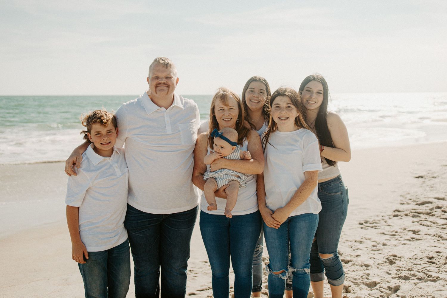 Family of six in matching white tops and jeans posing together on a sandy beach.