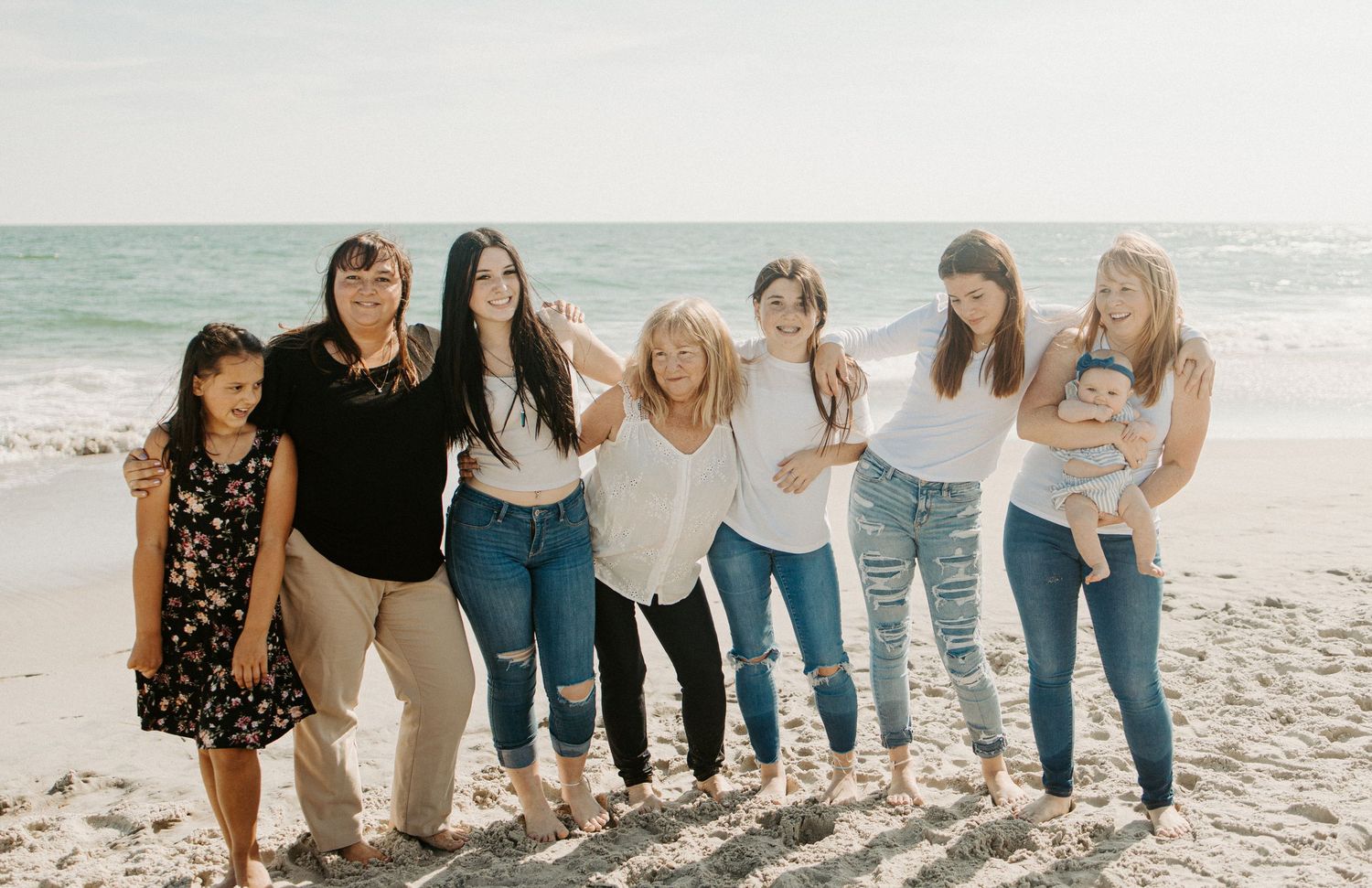 Group of seven women and girls posing together on a sunny beach with ocean backdrop.