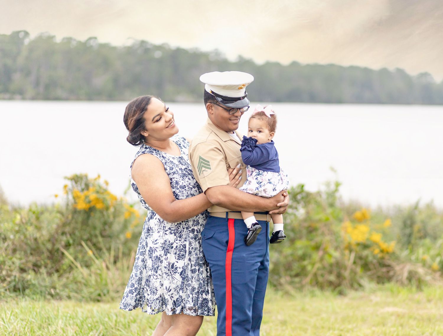 Marine in dress uniform holding baby with wife beside a scenic lakeside backdrop.