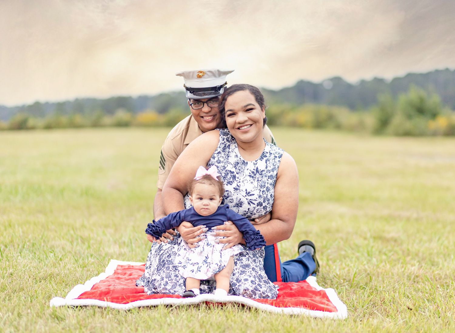 Marine in uniform sitting with wife and baby on a red blanket in a green field.