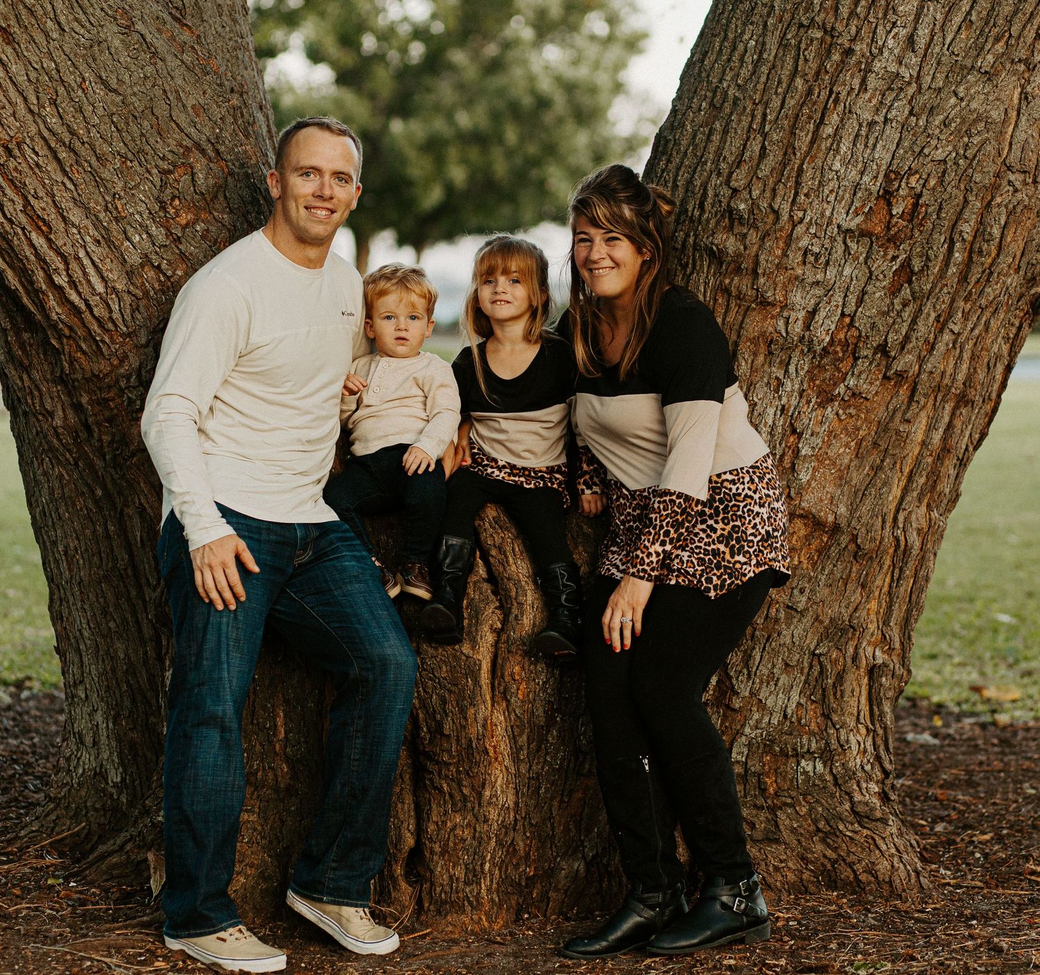 Family of four posing together against a large tree trunk in a park during a fall family portrait session.