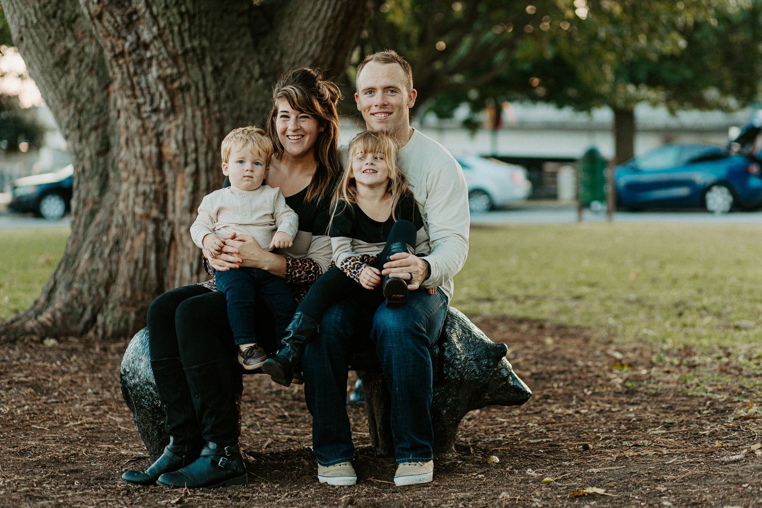 Family of four sitting on a bench under a large tree during an outdoor fall family portrait session.