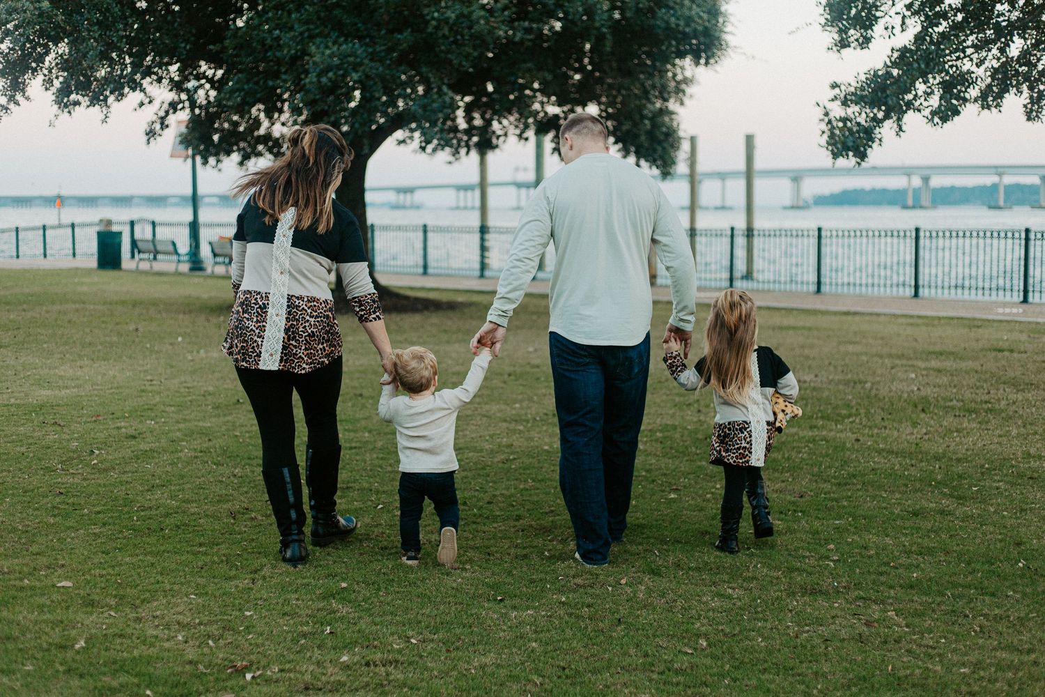 Family of four walking hand-in-hand across a grassy waterfront park during an outdoor family photo session.