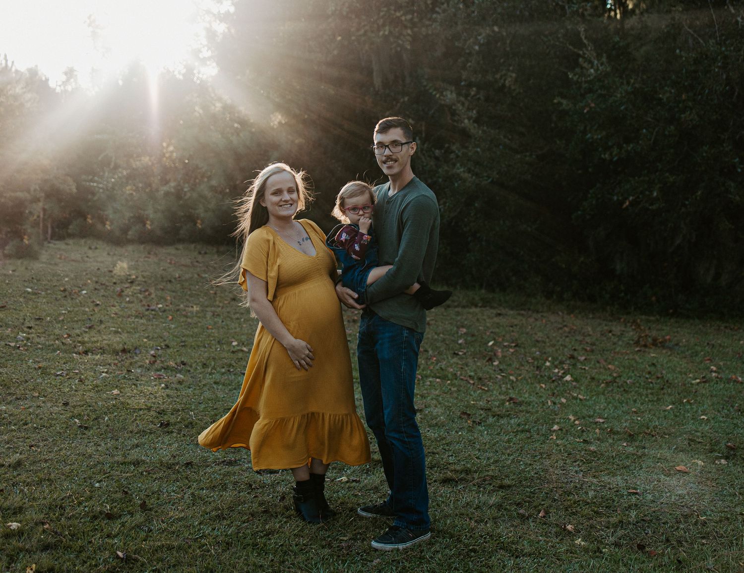 Happy expecting family with toddler posing in golden sunlight in a green meadow with trees behind them.