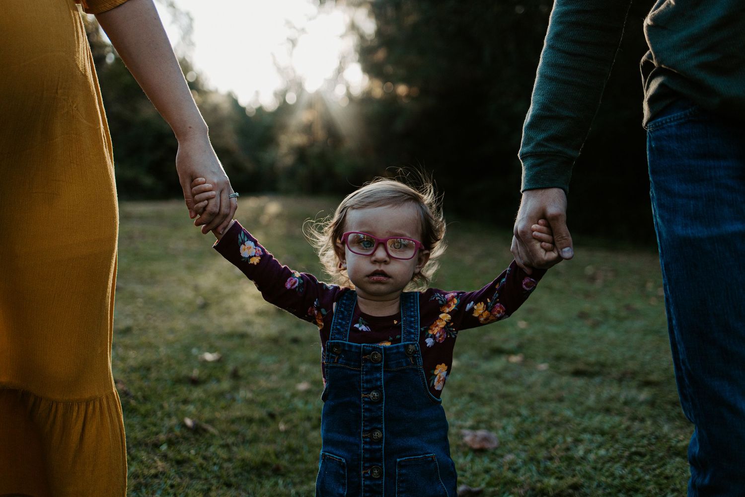 Adorable toddler in glasses holding parents hands while walking in a sunlit grassy outdoor setting.